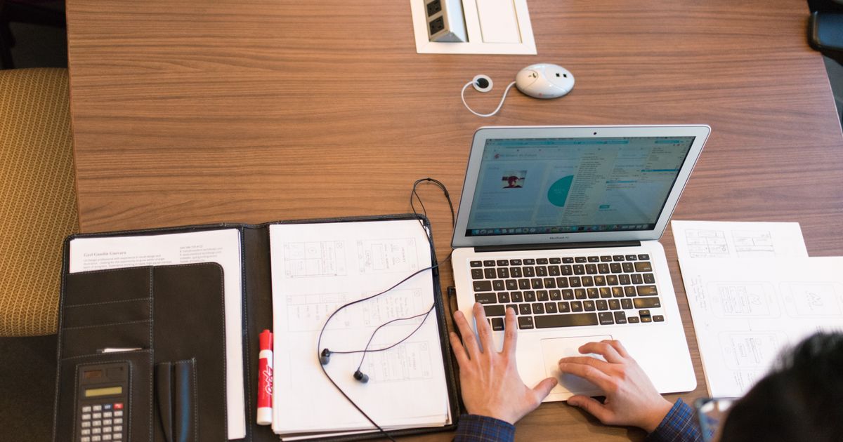 An office worker using a laptop alongside a notebook and printed documents.
