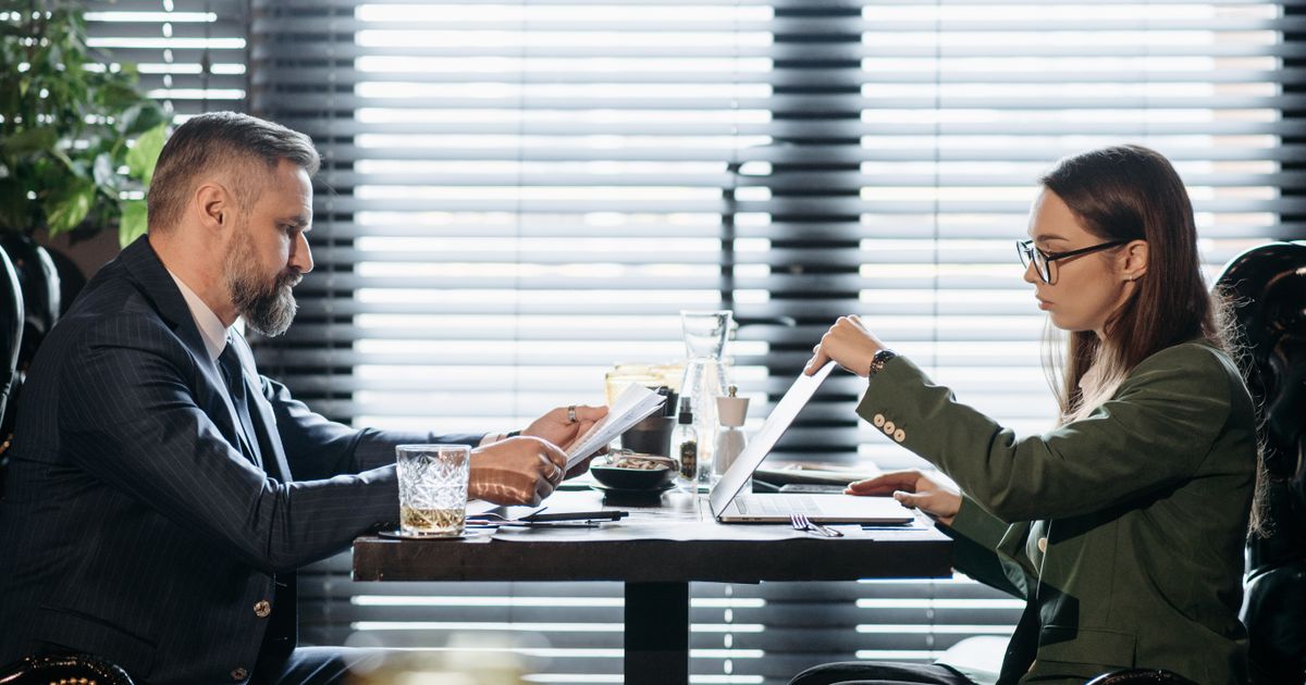 Two professionals reviewing documents across a meeting table with an open laptop.